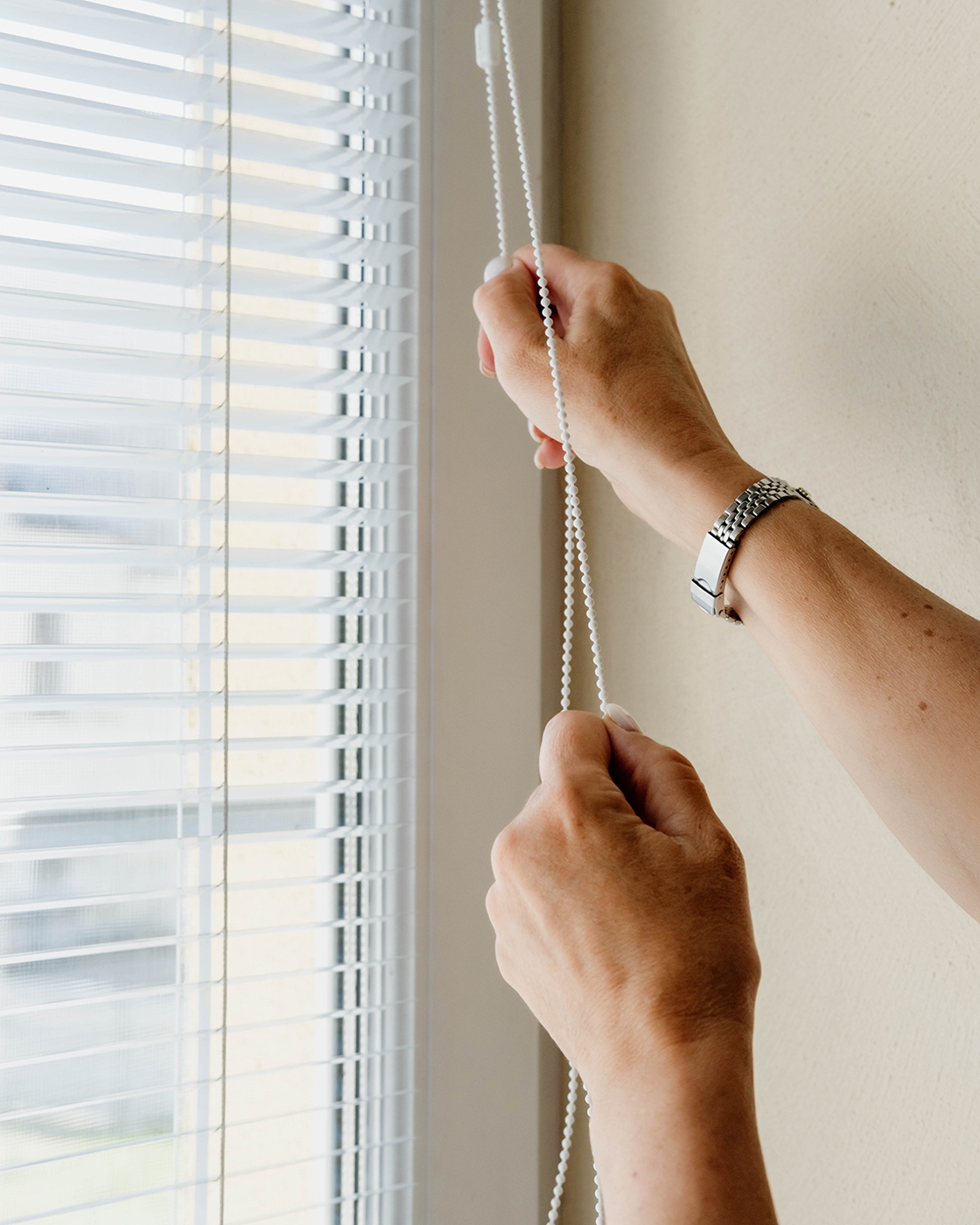 The woman opens and closes the horizontal white blinds with her hands.
